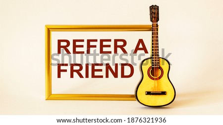 The text Refer a friend is written on a photo frame, next to a guitar on a white background. Referral marketing concept.