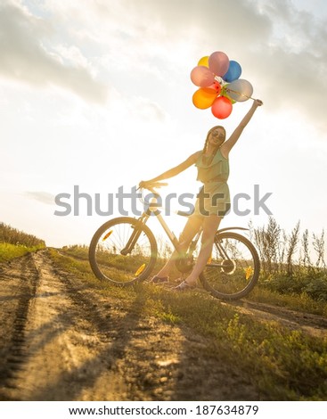 Portrait of Young Girl in dress with sunglasses riding bicycle flying air balloons on leash yellow sun set background Copy space for inscription Symbol of easy birthday celebration peace tranquility