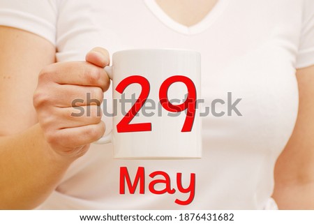 A female hand holds white mug with the red inscription May 29. Business and celebration concept