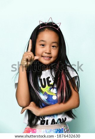 Close up of asian brunette girl in crown hoop and with colored strands in hair raises finger up having idea. Concept of children's style, positive emotions and adventures.
