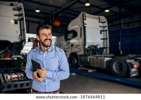 Smiling chief standing in auto park and holding tablet. In background are trucks. Firm for import and export. Royalty-Free Stock Photo #1876464811