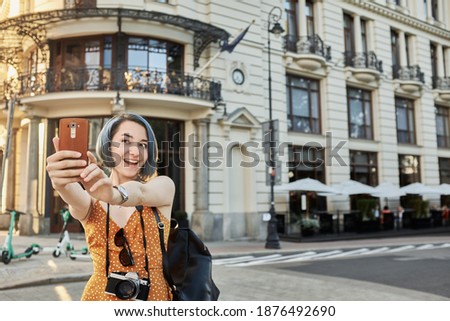Cute girl in orange dress with blue hair and camera on her neck takes a selfie against the background of old European architecture