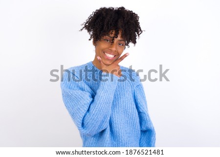 Young beautiful African American woman wearing blue knitted sweater against white wall, looking confident at the camera smiling with crossed arms and hand raised on chin. Thinking positive.