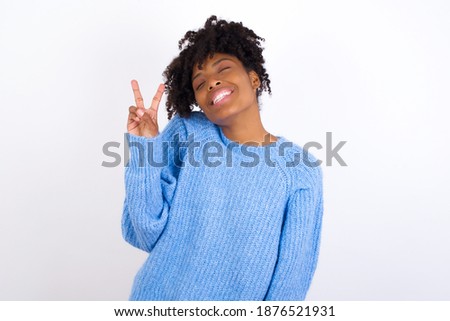 Young beautiful African American woman wearing blue knitted sweater against white wall, smiling with happy face winking at the camera doing victory sign. Number two.