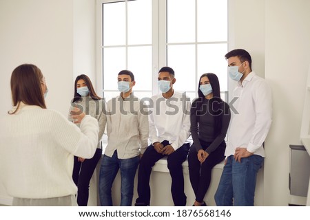 People wearing protective medical face mask in company meeting. Diverse audience listening to business speaker. Coach talking to team of students or office workers, sharing knowledge and giving advice