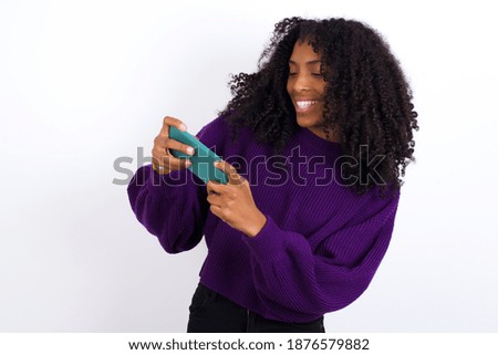 Young beautiful African American woman wearing purple knitted sweater against white wall, holding in hands cell playing video games or chatting