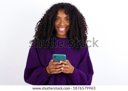 Young beautiful African American woman wearing purple knitted sweater against white wall, enjoys distant communication, uses mobile phone, surfs fast unlimited internet, has pleasant smile.