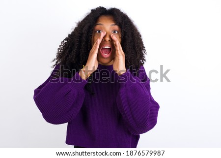 Young beautiful African American woman wearing purple knitted sweater against white wall, shouting excited to front.