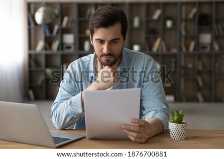 Pensive young Caucasian man sit at desk at home work on laptop read paper document. Thoughtful millennial male distracted from computer job consider post paperwork or letter correspondence news. Royalty-Free Stock Photo #1876700881