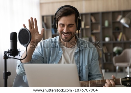Happy millennial male artist in headphones and guitar have online video music lesson on computer. Smiling young man singer tutor or coach talk greet on webcam digital virtual training at home studio. Royalty-Free Stock Photo #1876707904