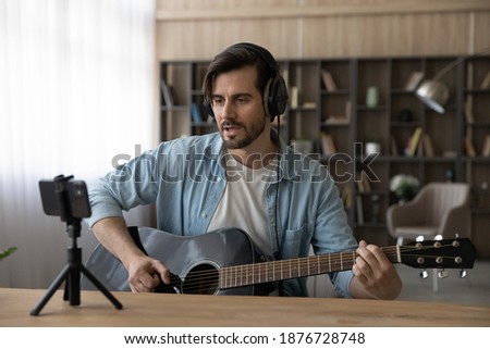 Young Caucasian male singer in headphones hold play guitar record new single on smartphone at home studio. Millennial man artist use musical instrument sing shoot music video on cellphone camera. Royalty-Free Stock Photo #1876728748