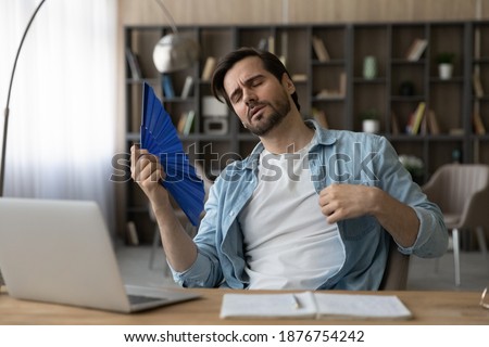 Exhausted millennial Caucasian male employee sit at desk at home office feel overheated use waver. Tired young man breathe fresh air wave with hand fan, suffer from hot weather or lack or AC. Royalty-Free Stock Photo #1876754242