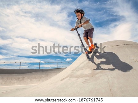 Child wearing a helmet goes down a ramp with a scooter in a skate park on a sunny summer day Royalty-Free Stock Photo #1876761745