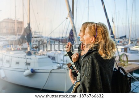 Portrait of beautiful mature mother and her daughter making a selfie using smart phone and smiling at the seaport with sailing ship in background
