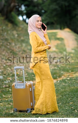 Gorgeous smiling muslim woman in traditional wear holding smartphone and  luggage at lake garden.