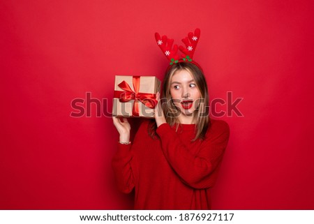 Amazed young Santa girl in fun decorative deer horns on head pointing index finger on red boxes with gifts presents isolated on red background. Happy New Year 2019 celebration holiday party concept