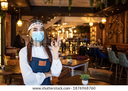 Portrait of a beautiful happy waitress working at a restaurant wearing a facemask to avoid the spread of coronavirus and showing OK sign. Food service concepts