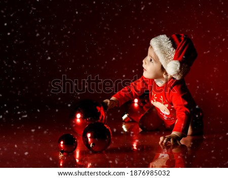 Infant baby girl in red bodysuit and Santa hat sits on her knees surrounded with Christmas New Year tree balls touching one and looks at copy space upper corner under snow over dark background.