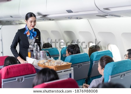 Asian female flight attendant serving food and drink to passengers on airplane. The cabin crew pushing the cart on aisle to serve the customer. Airline service job and occupation concept. Royalty-Free Stock Photo #1876988008