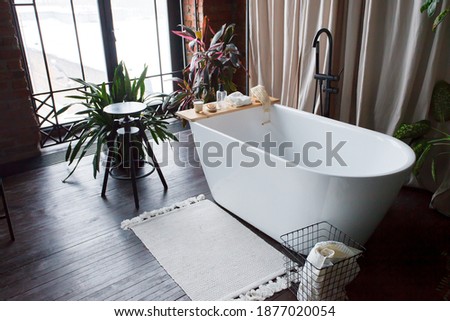Modern bathroom with freestanding white acrylic bathtub and black loft mixer. Walls made of bricks, big window, parquet is wooden on floor Royalty-Free Stock Photo #1877020054