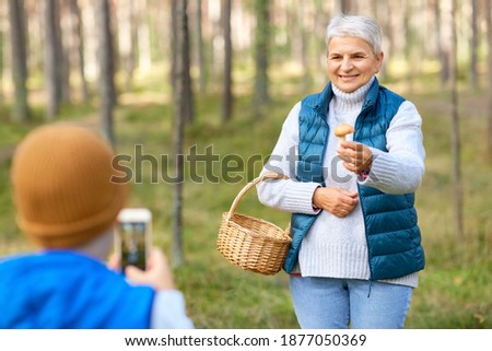 picking season, leisure and people concept - grandson with smartphone photographing happy smiling grandmother with mushroom and basket in forest