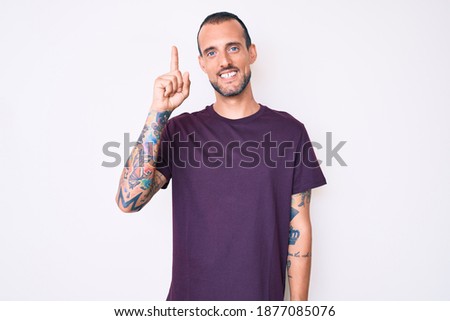 Young handsome man with tattoo wearing casual clothes showing and pointing up with finger number one while smiling confident and happy. 