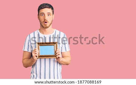 Handsome young man with bear holding empty frame scared and amazed with open mouth for surprise, disbelief face 
