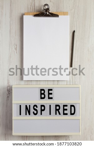 'Be inspired' on a lightbox, cliboard with blank sheet of paper on a white wooden surface, top view. Flat lay, overhead, from above. 