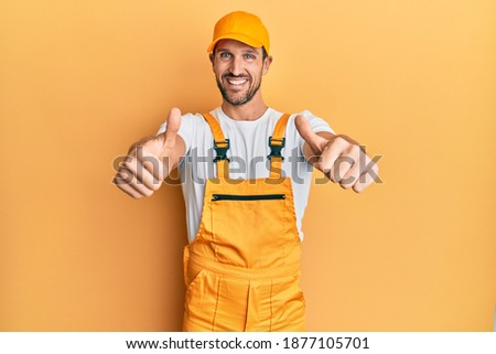 Young handsome man wearing handyman uniform over yellow background approving doing positive gesture with hand, thumbs up smiling and happy for success. winner gesture. 