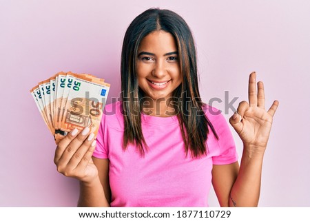 Young latin girl holding euro banknotes doing ok sign with fingers, smiling friendly gesturing excellent symbol 