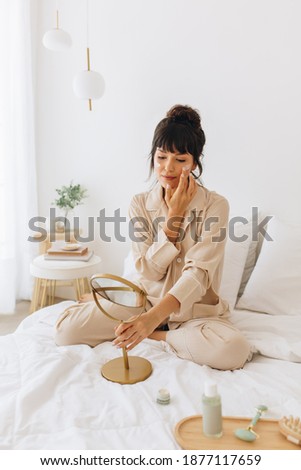 Woman doing beauty self care sitting at home. Woman sitting on bed applying cosmetics looking at small mirror. Royalty-Free Stock Photo #1877117659