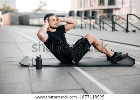 Fit man pumping press lying on black yoga mat. Muscular man doing his favorite morning exercise. Black sport clothes. Fit happy man doing fitness exercises outdoors. Street on background. Royalty-Free Stock Photo #1877118595