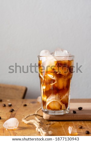 Cold refreshing coffee with ice and creamy clouds in a glass on a wooden board close-up, cocoa beans are scattered on the table