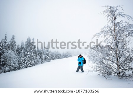 Young male traveler with backpack takes pictures of beautiful tall snowy fir tree in a high snowdrift against the backdrop of fog on a frosty winter day. Trekking and travel concept. Advertising space