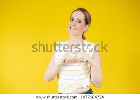 Young woman in casual t-shirt making heart figure over yellow background.