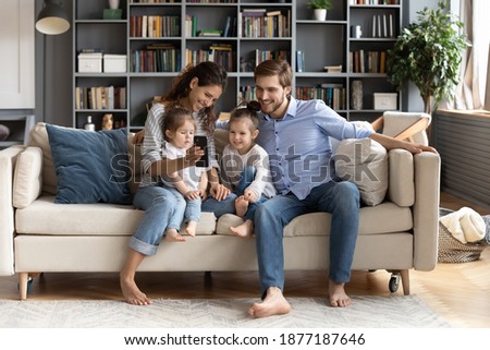 Happy young Caucasian family with two small daughters sit relax on couch in living room watch funny video. Smiling parents rest on sofa at home use talk on call on modern smartphone.