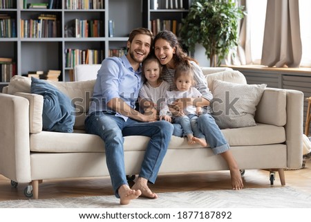 Portrait of overjoyed young Caucasian family with two little daughters sit rest on cozy couch in living room. Happy parents and small girls children rest on comfortable sofa enjoy weekend at home.