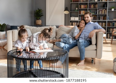 Loving young Caucasian parents relax on couch in living room, two little daughters paint draw on table. Happy family with small girls children rest at home, enjoy leisure weekend activity together.