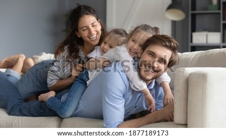 Wide banner panoramic portrait of happy young Caucasian family with two small daughters relax on couch at home. Smiling parents rest on sofa in living room together with little girls children.
