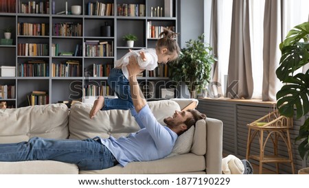 Wide banner panoramic view of smiling young dad rest on sofa at home hold throw in air little daughter. Happy loving father feel playful have fun play with small girl child, relax together on couch.
