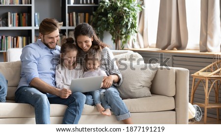 Wide banner panoramic view of happy young Caucasian family renters with two small daughters relax on sofa using laptop together. Smiling parents with little girls children browse internet on computer.