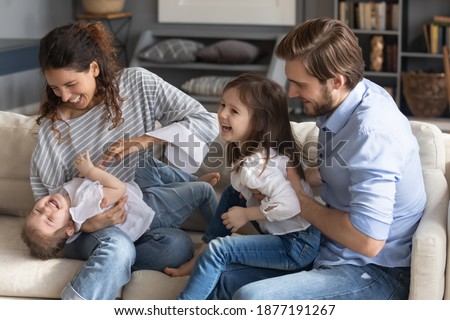 Overjoyed young Caucasian family sit on sofa in living room play tickle little daughters. Happy mother and father feel playful have fun with two small girls children, enjoy weekend at home together. Royalty-Free Stock Photo #1877191267