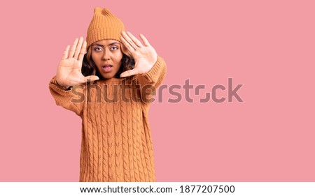 Young beautiful mixed race woman wearing wool sweater and winter hat doing frame using hands palms and fingers, camera perspective 