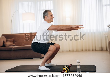 Workout time Strong senior man doing squat exercises at home in his light living room. Healthy lifestyle, wellbeing and activity concept. Black and white sportswear. Active and happy old age. Royalty-Free Stock Photo #1877232046