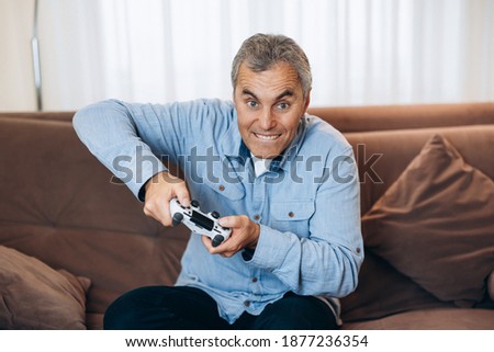 Impossible to put down Mature man is completely absorbed in playing his favorite game. He presses hard on buttons on game console. Living room on background. Male gamer having a good time.