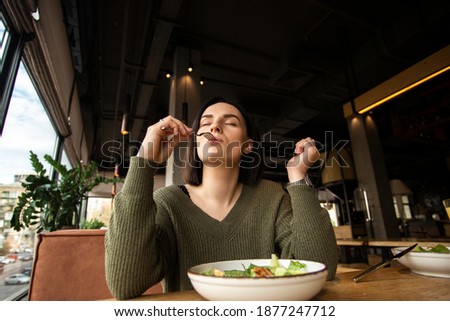 Satisfied young woman enjoys tasty salad in a restaurant, putting fork into her mouth and closing her eyes. Good customer service. Healthy diet concept. Weight loss without struggle. Royalty-Free Stock Photo #1877247712