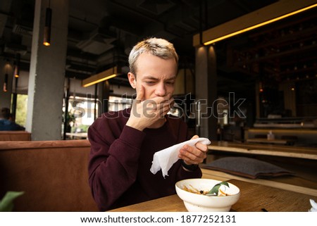 Spoiled food in the restaurant. Dissatisfied young man in a cafe covers his mouth with a hand disgusted with a taste of food. A meal on the table in front of him. Bad customer service concept. Royalty-Free Stock Photo #1877252131