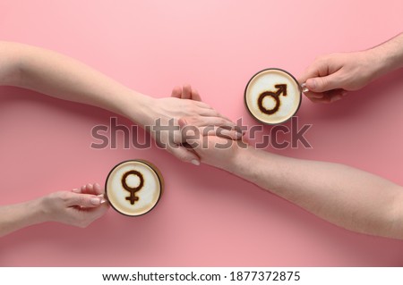 Two cups of coffee with symbols of venus and mars on milk foam and holding hands of couple in love on pastel pink background. Concept romantic date on Valentine's day. Top view, creative flat lay