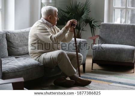 Bones ache on weather. Thoughtful mature male with grey hair resting on sofa leaning on stick feeling hard to move. Sad tired old man suffer of walking with limp need rehabilitation after leg trauma Royalty-Free Stock Photo #1877386825