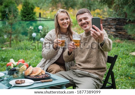 Video conference with friends in the open air during a picnic in nature with beer and barbecue. Young people use their smartphone to chat with friends and have fun